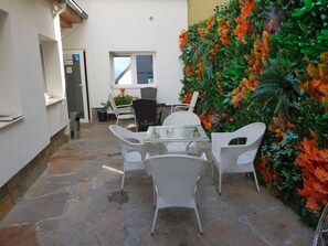 Une terrasse dotée de meubles en osier et d’une table en verre, entourée d’un mur décoré de feuillage artificiel.