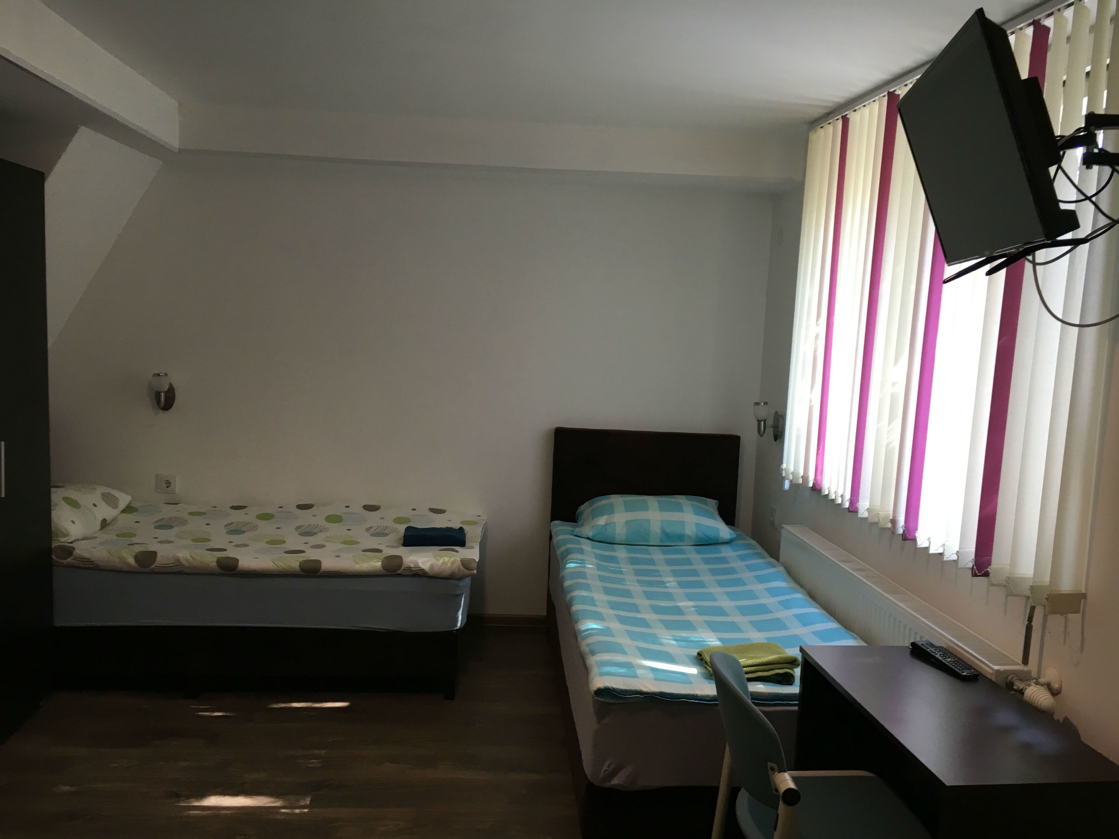 A small, well-lit room with two beds, a desk, a chair, and a television mounted on the wall.