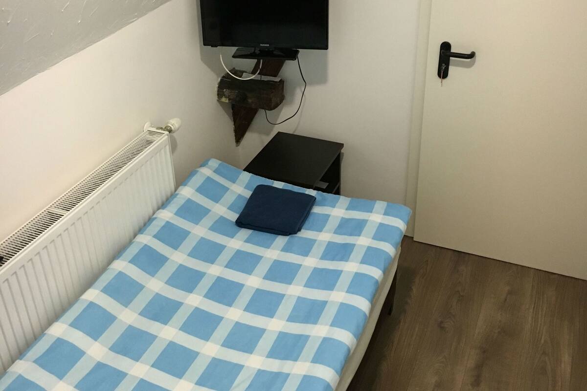 Una habitación pequeña y funcional con una cama individual, televisor montado, radiador y puerta.