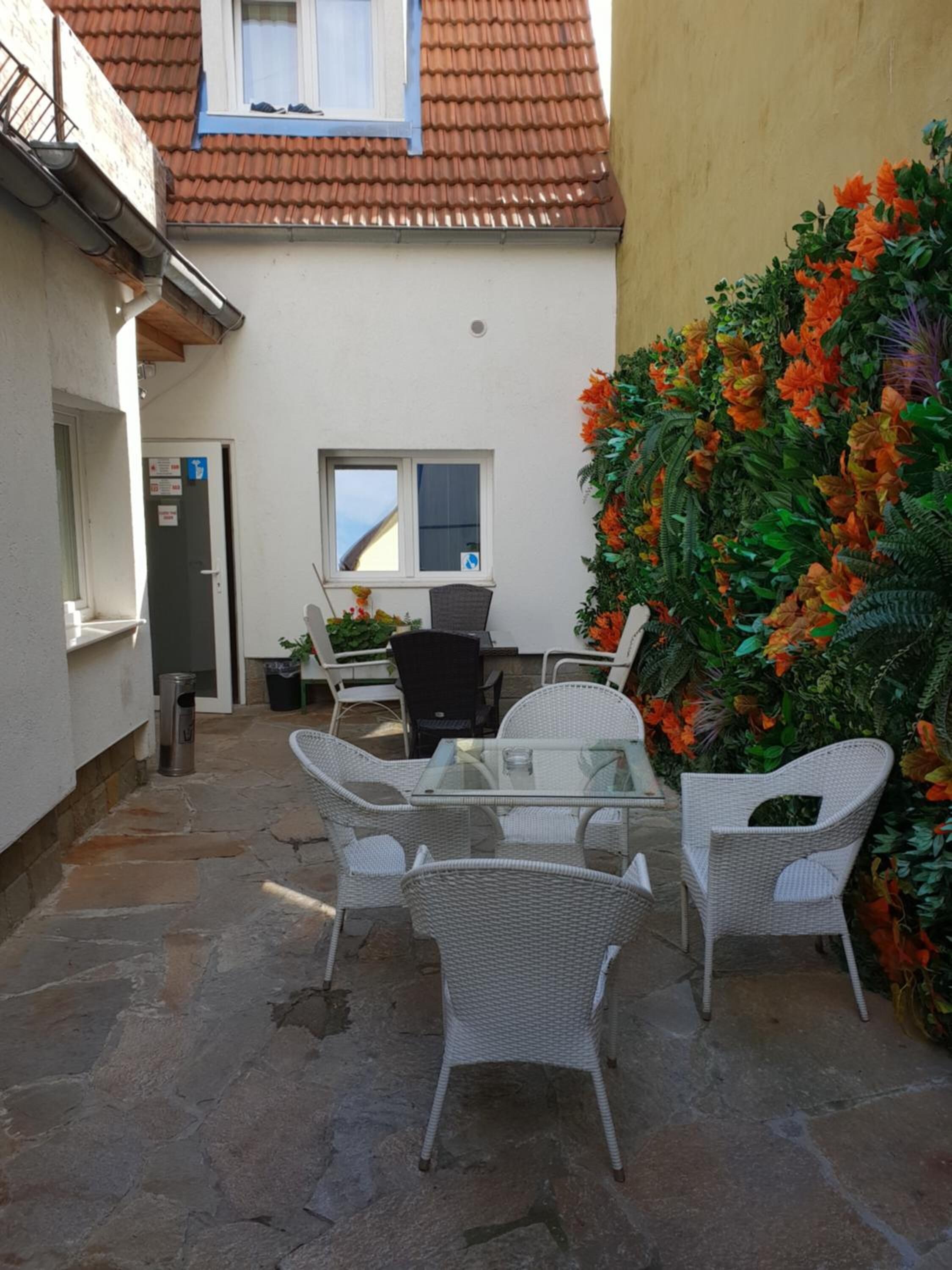 Un pequeño patio al aire libre con una mesa de vidrio y sillas de mimbre, una ventana con vistas y una pared decorada con plantas artificiales.