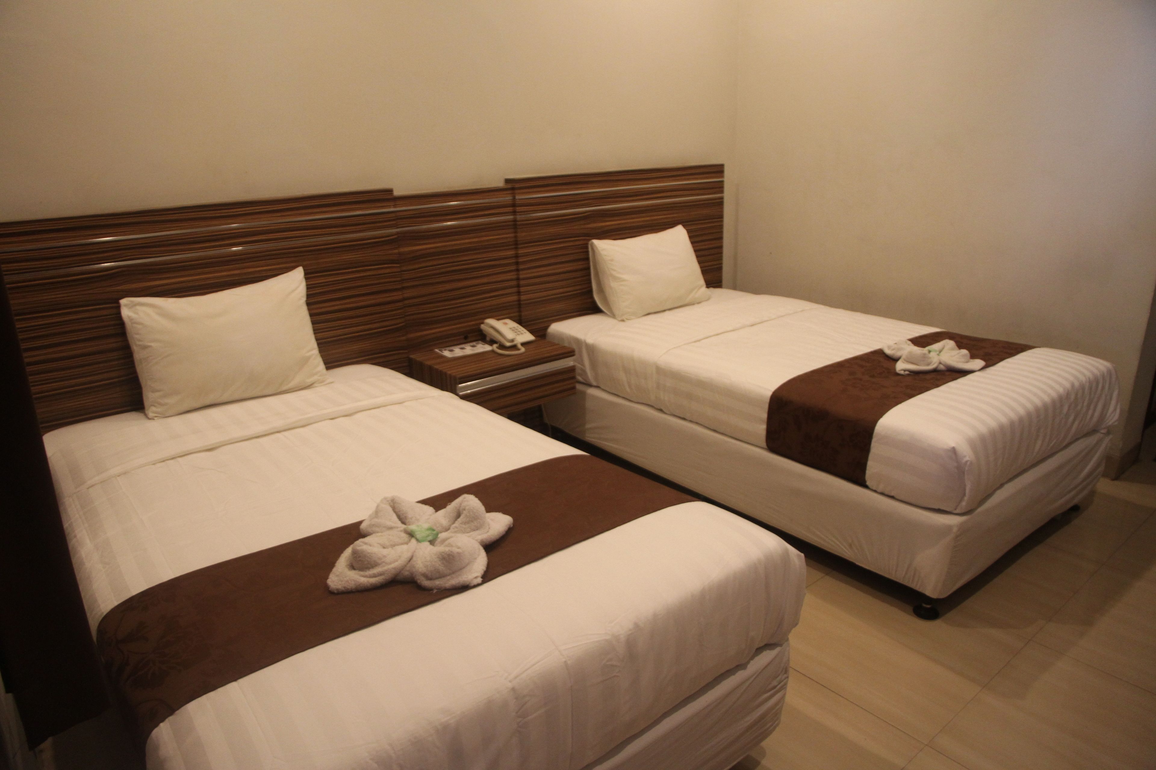 A hotel room with two single beds, each with a wooden headboard, white bedding, and a brown accent on the bedspread. There is a bedside table with a phone and a small lamp.