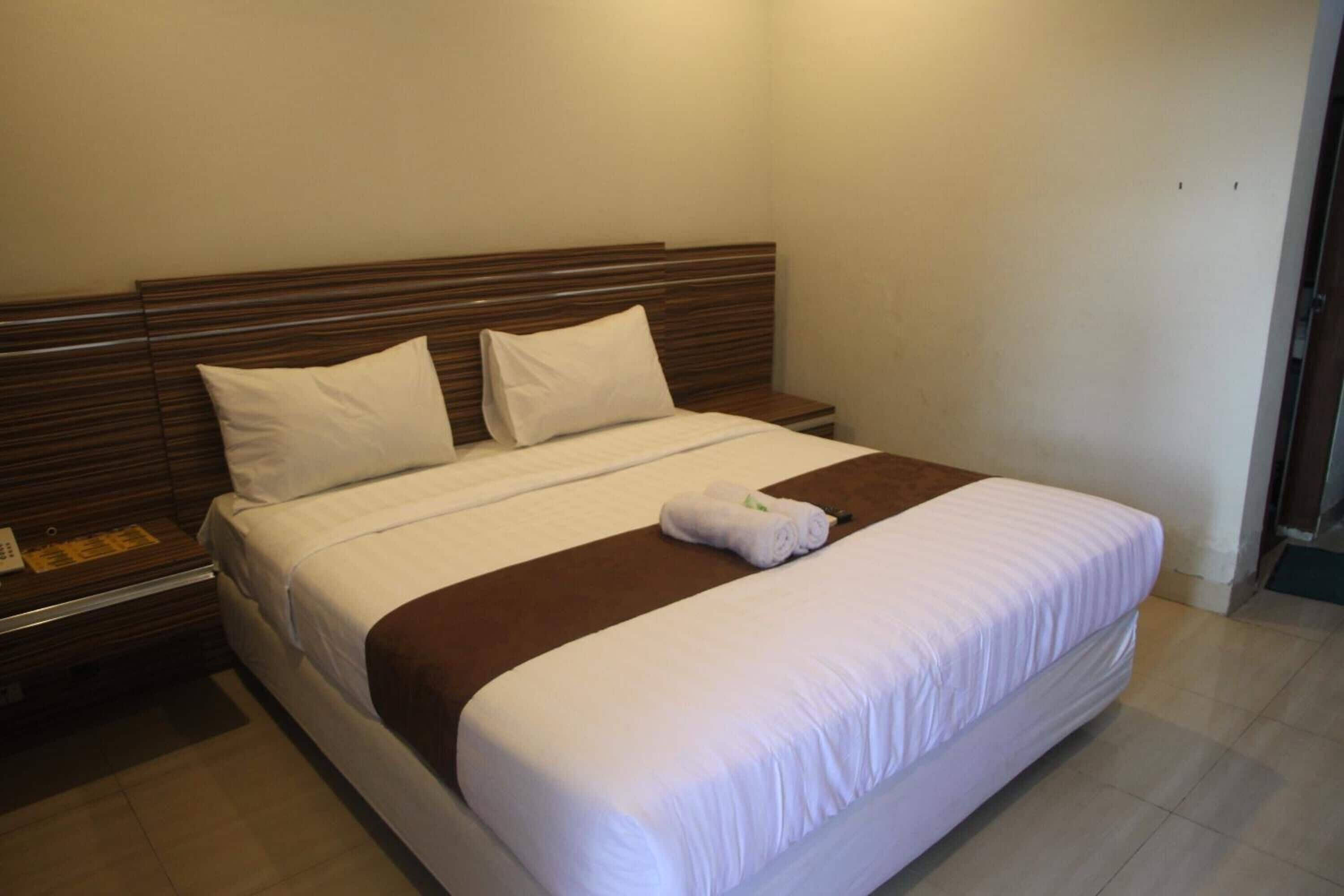 A neatly made bed with white linens and a brown accent, a wooden headboard, and a nightstand with a lamp and a small table.