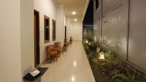 A hallway with wooden chairs and tables, a trash bin, and a small garden area with plants and a lantern. - Palapa Hotel (Mataram)