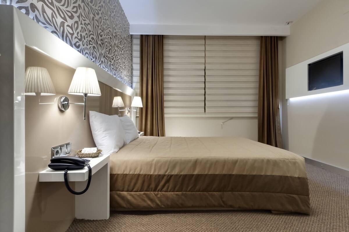 A hotel room with a large bed, two bedside lamps, a telephone, and a flat-screen TV mounted on the wall.