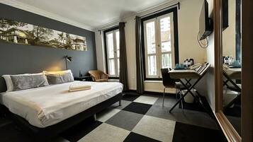 Habitación de hotel con una cama, un escritorio y una silla. Cuenta con una ventana con cortinas, un cuadro en la pared y un piso a cuadros.