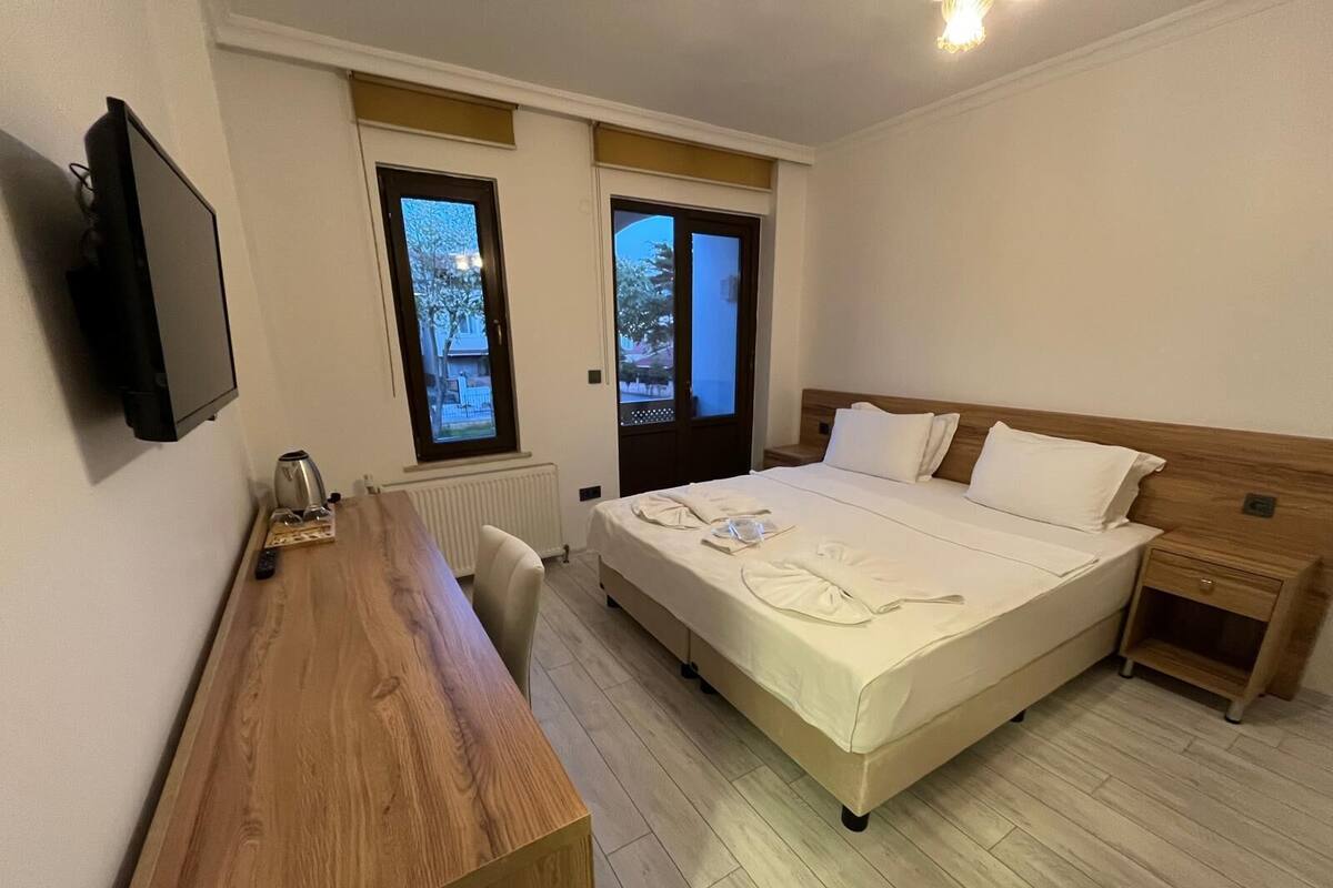 A hotel room with a wooden desk, a bed with white linens, a television on the wall, and two windows with curtains.