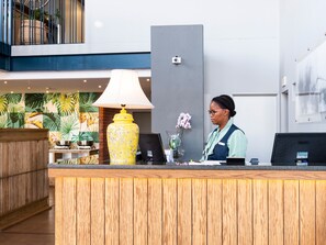 A reception desk with a wooden counter, a yellow vase with flowers, a lamp, and a modern wall with a security camera. - Urban Hotel Kathu (Kathu)