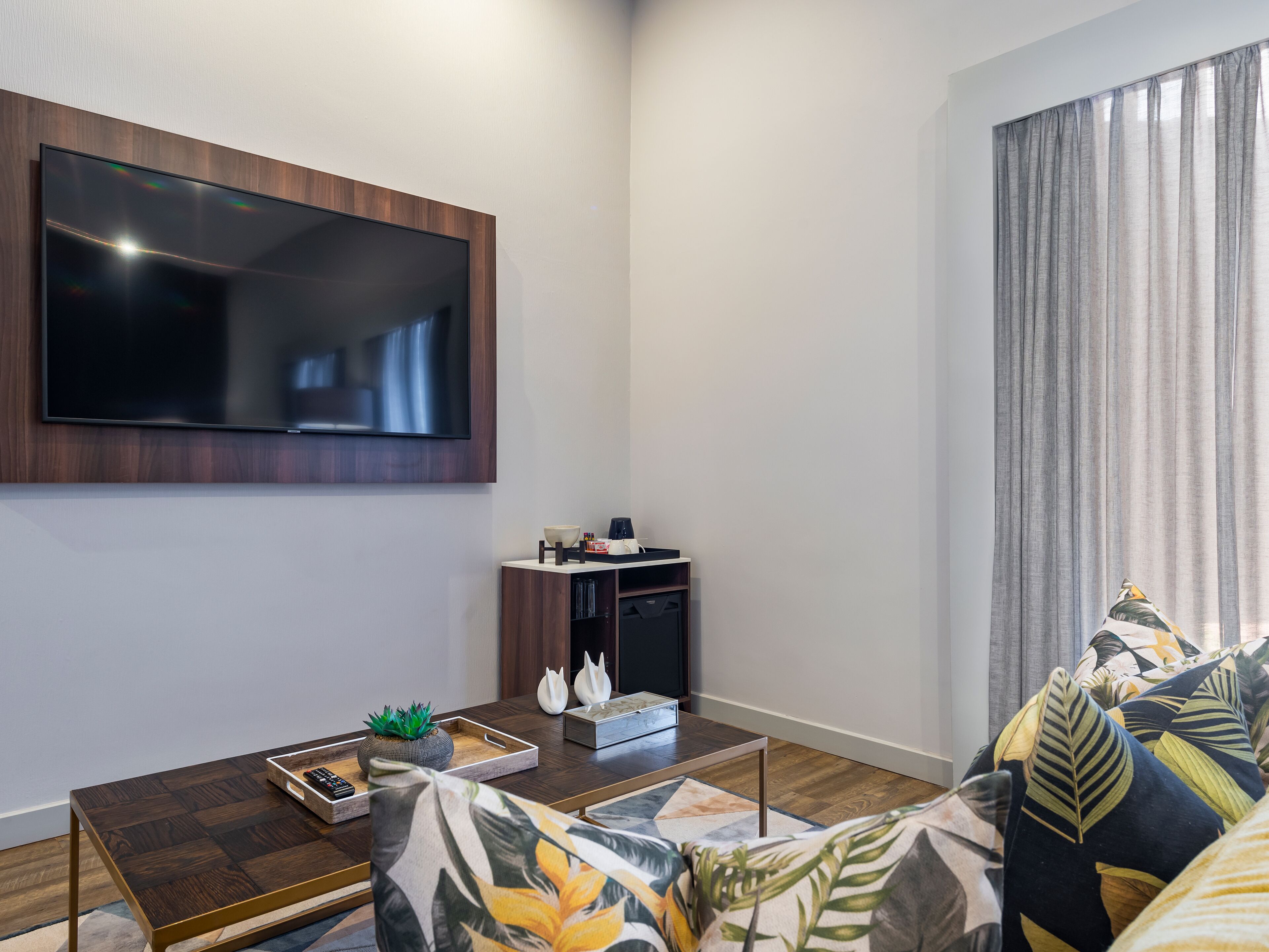 A modern living room with a flat-screen TV mounted on the wall, a coffee table with decorative items, a sofa with patterned cushions, and a side table with a plant.
