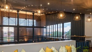 A modern interior with a long bench, multiple hanging light fixtures, and large windows offering a view of the outdoors. - Urban Hotel Kathu (Kathu)