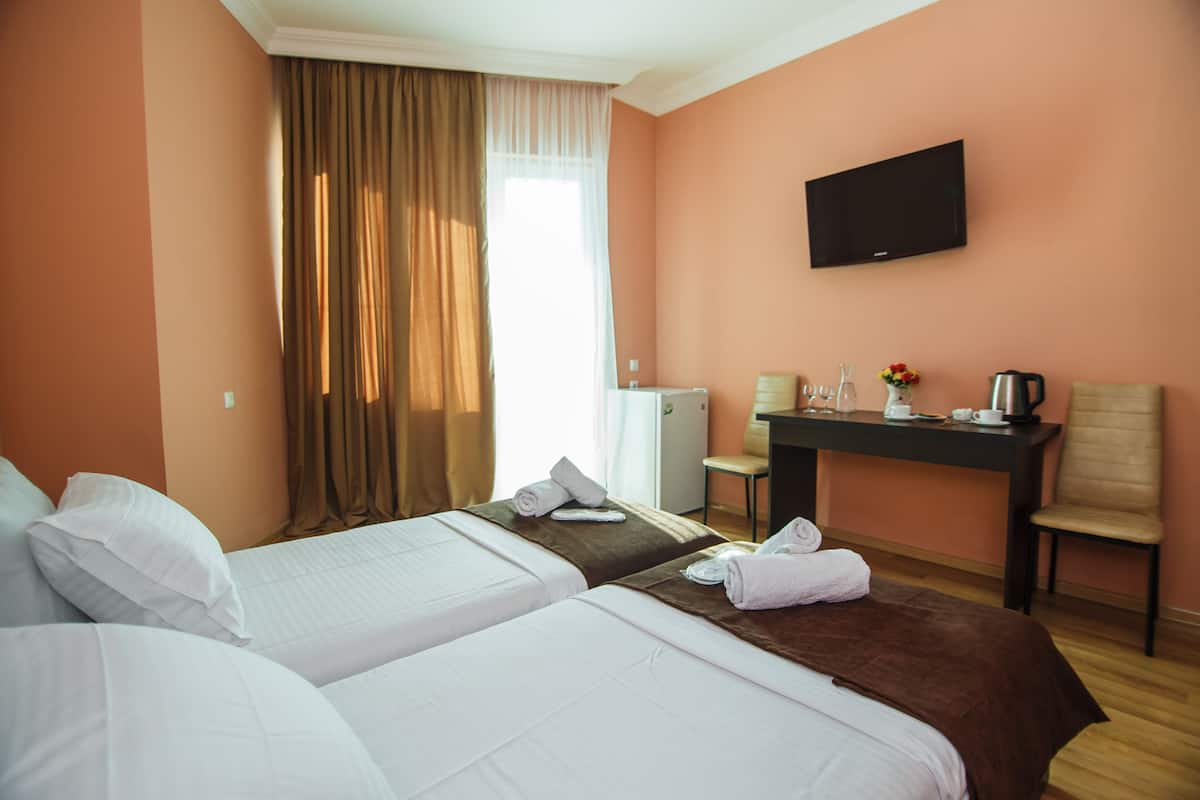 A hotel room with two beds, a desk with a flat-screen TV, a chair, and a small table with a kettle and cups.
