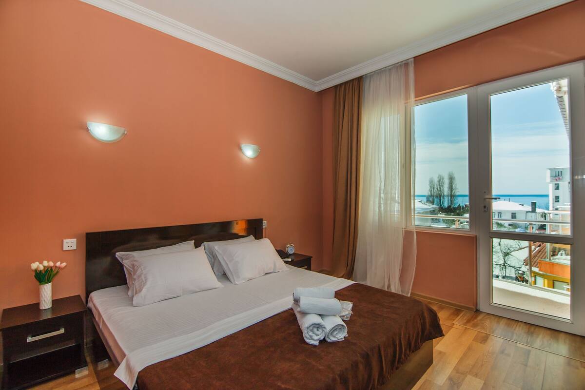 A hotel room with a bed, a nightstand, a vase with flowers, and a view of buildings through a large window.