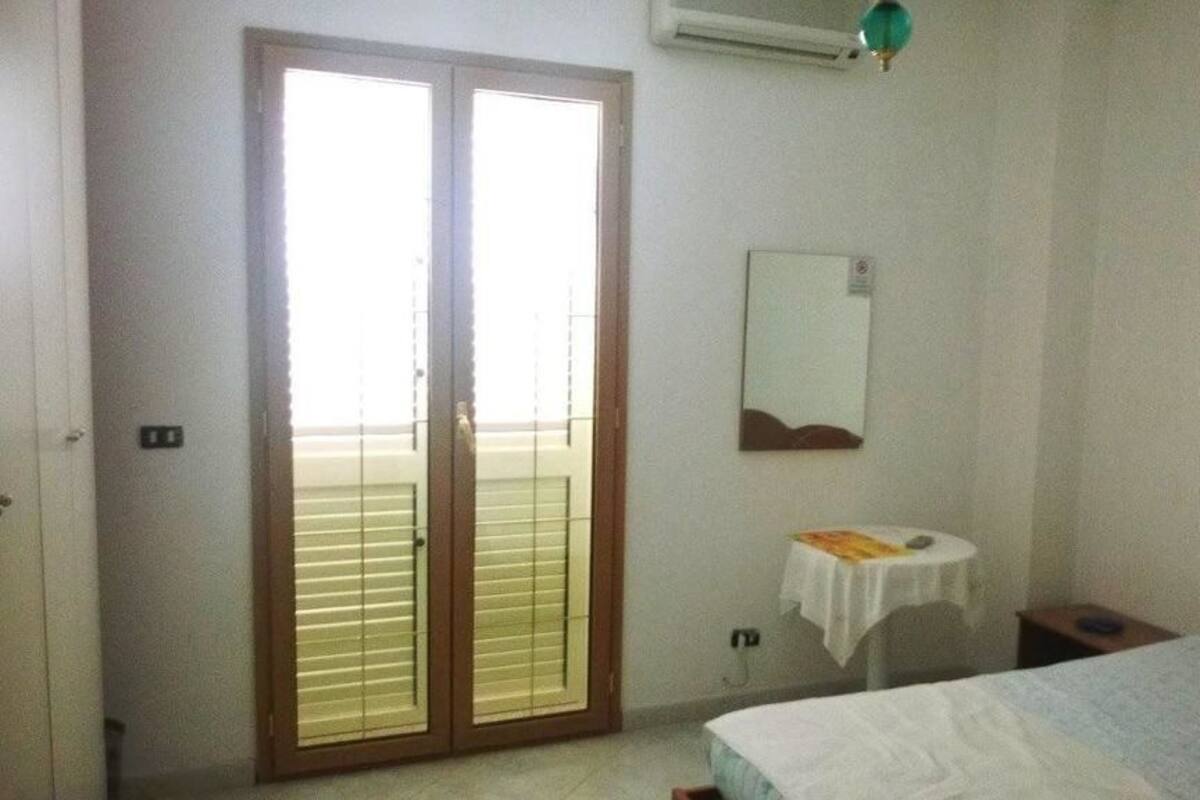 A bedroom with a bed, a small bedside table, a mirror, and a sliding door to a balcony.