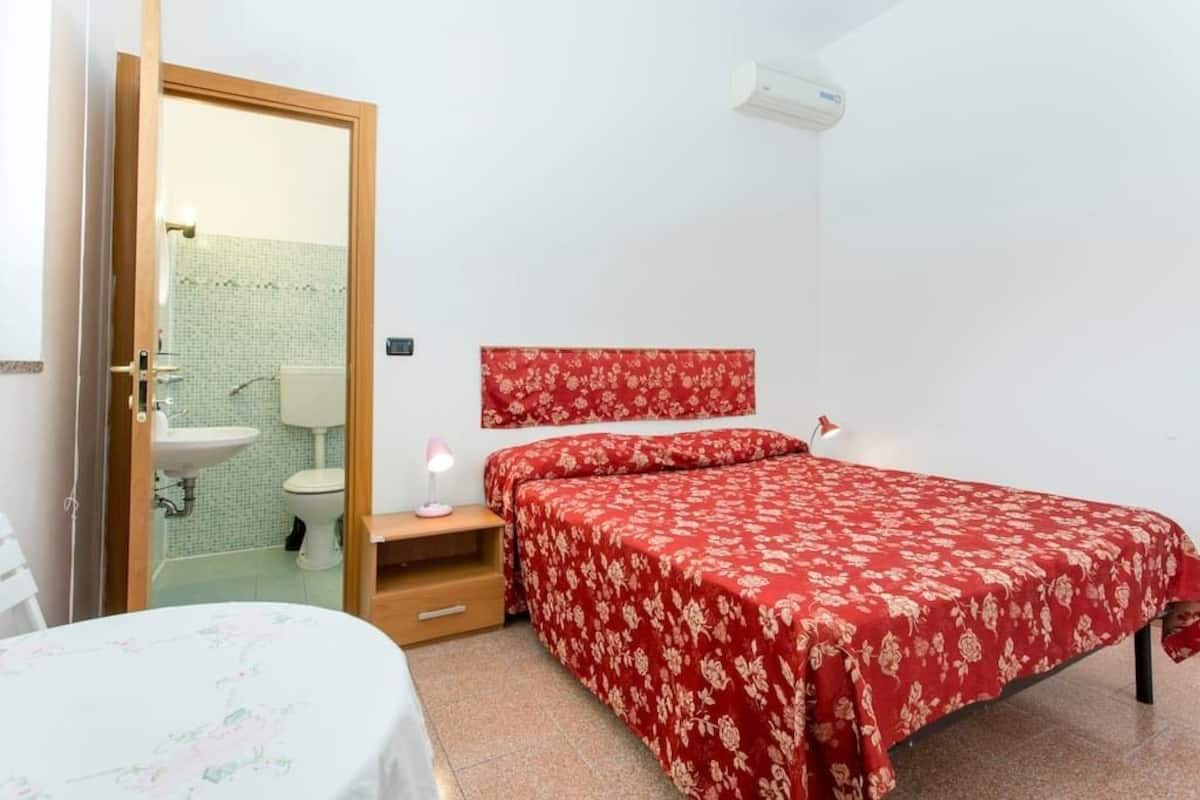 A bedroom with a bed, a bedside table, a nightstand, a mirror, a red floral headboard, and a bathroom visible through an open door.