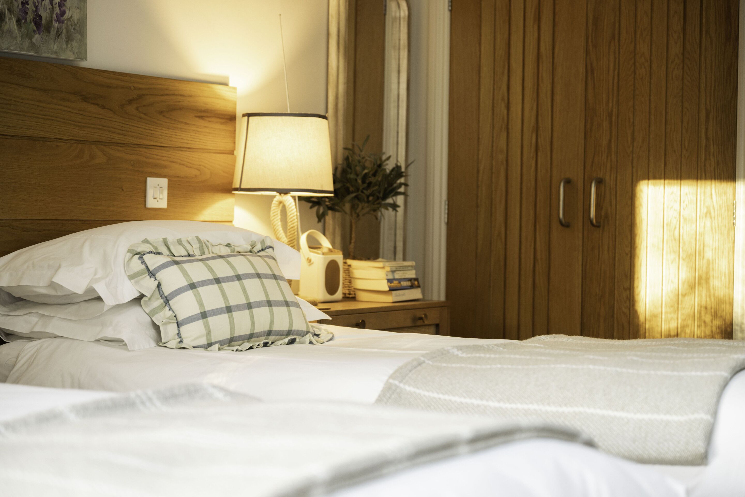 A neatly made bed with white linens and a plaid pillow, a bedside lamp, a small table with books and a radio, and a wooden wardrobe.