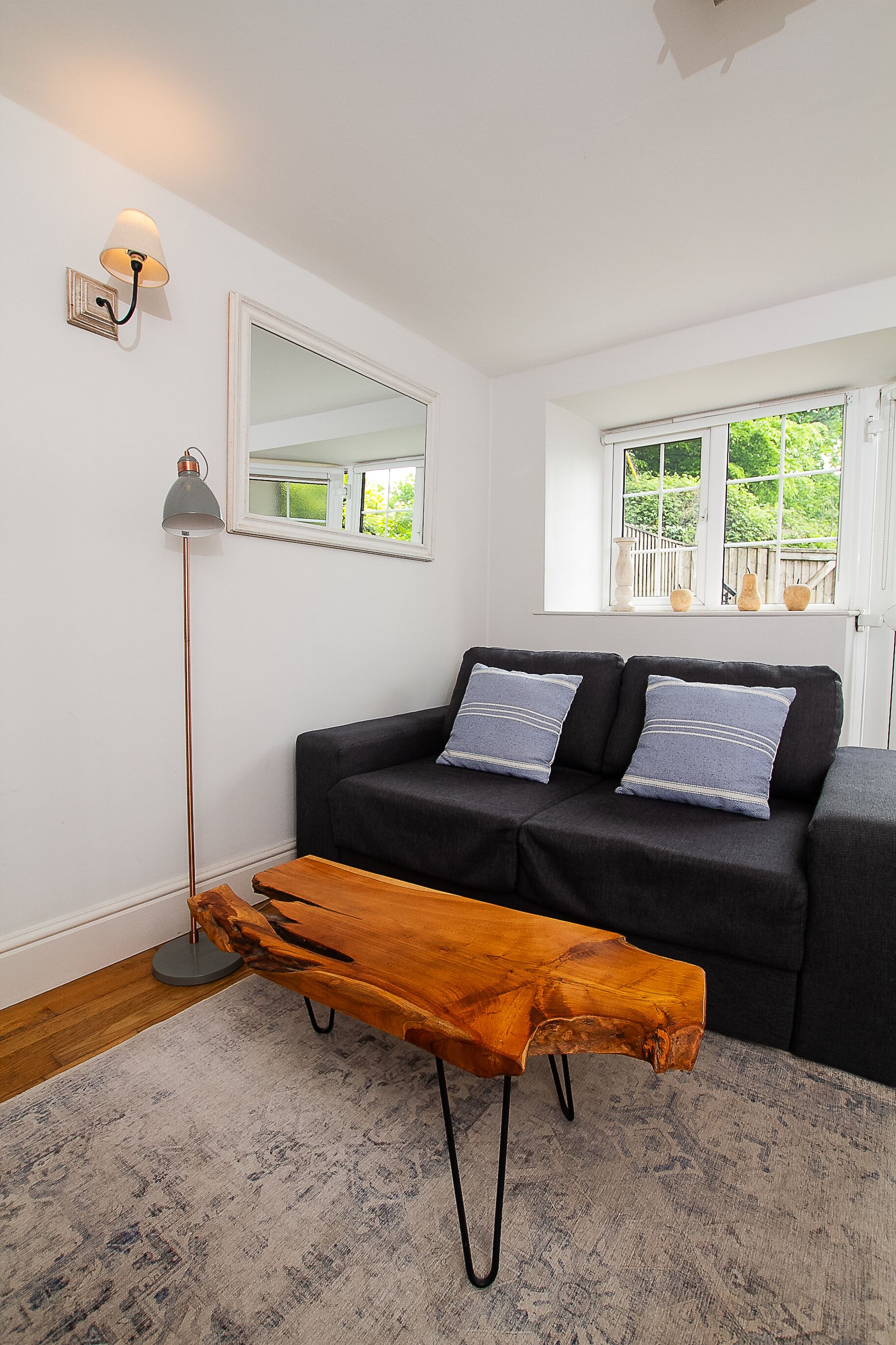 A living room with a dark sofa, two cushions, a wooden coffee table, a floor lamp, and a mirror on the wall.