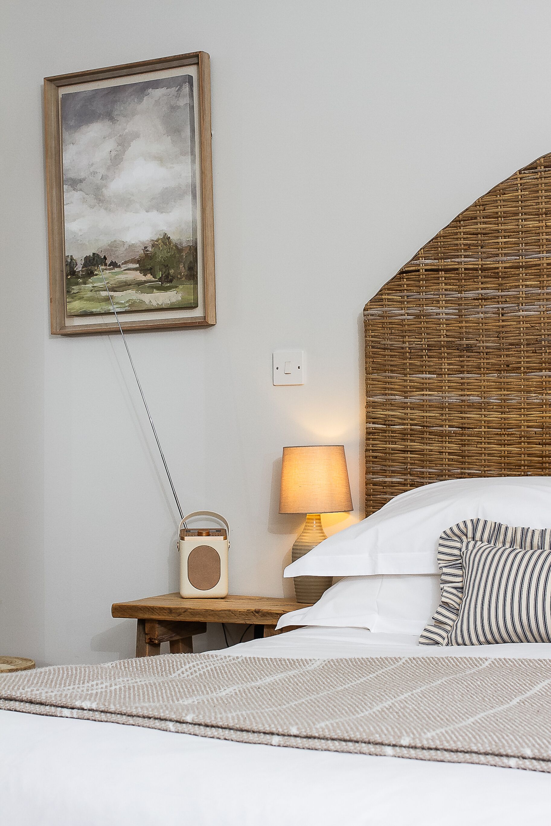 A neatly arranged bedroom with a bed, a bedside table with a lamp, a radio, and a framed painting on the wall.