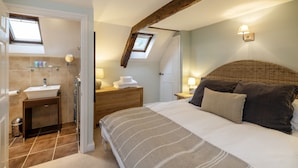 A bedroom with a large bed, a bedside table, a nightstand, a window, and a bathroom with a sink and mirror. - Winyard's Gap Inn (Beaminster)