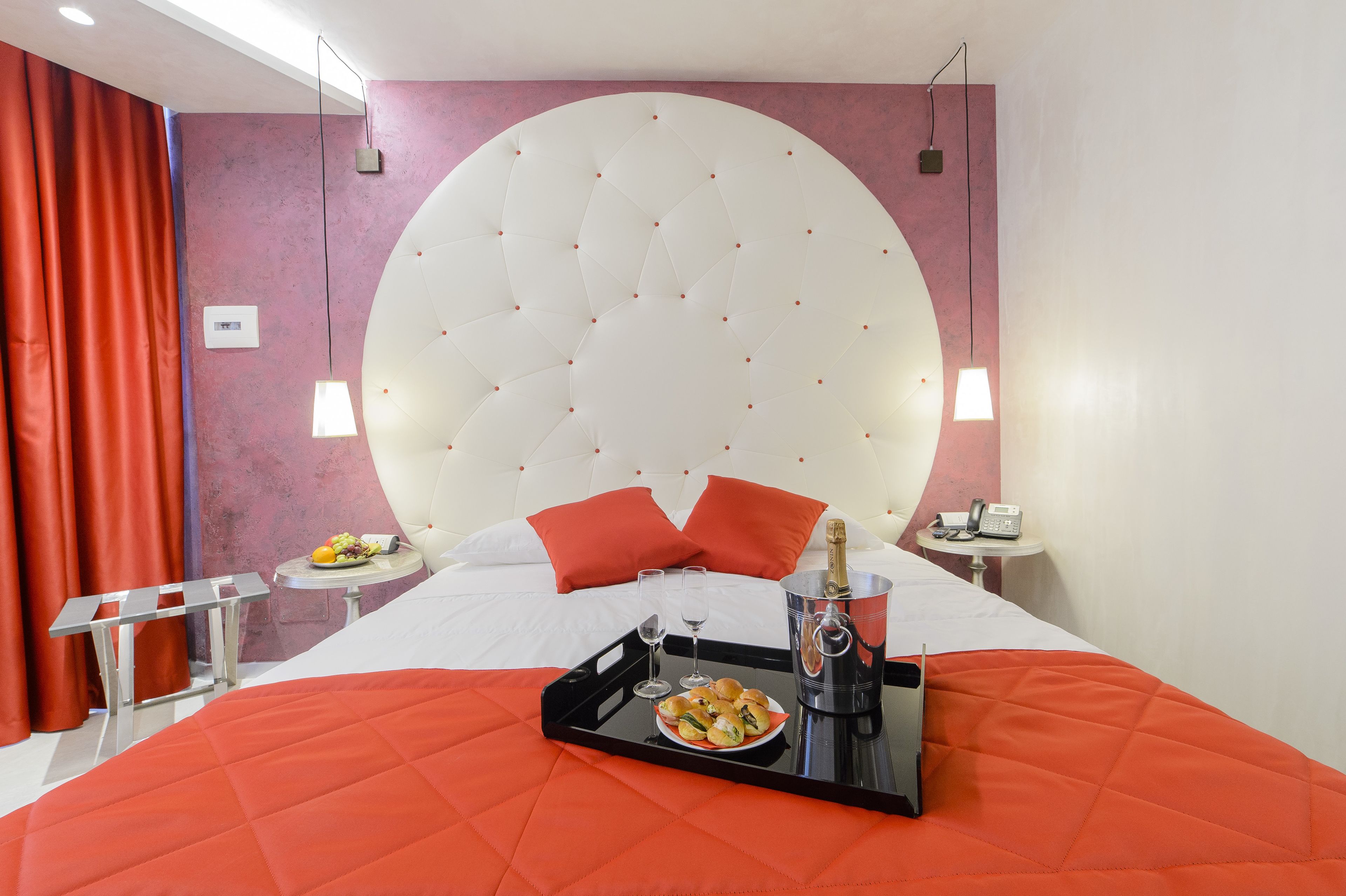 A hotel room with a large bed, a bedside table with a phone and a bottle, a tray with food and glasses, and a red curtain.