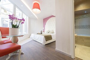 Et moderne hotelværelse med en seng, et glasbord med blomster, en rød stol og udsigt over byen gennem store vinduer.