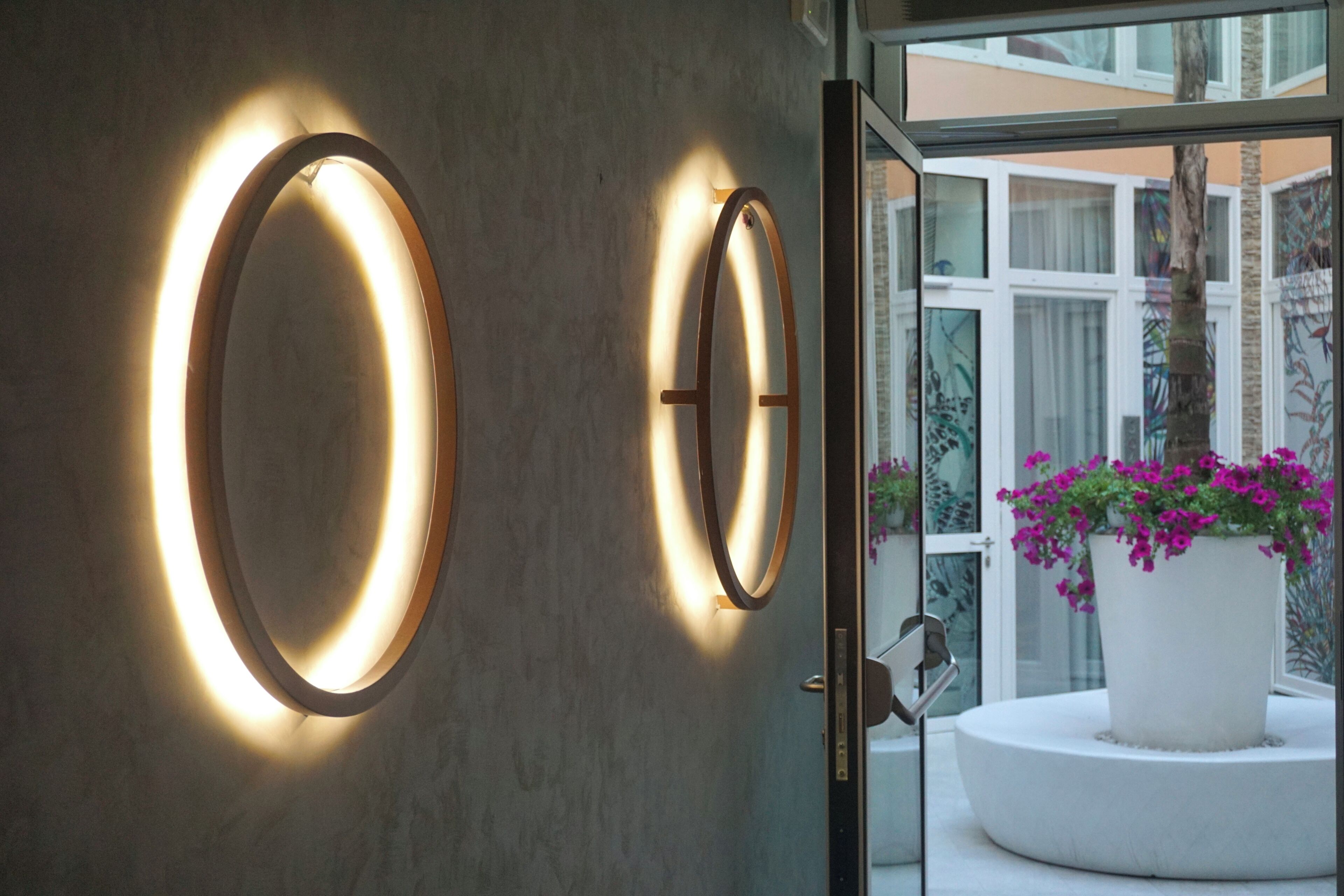 A modern interior with two illuminated wall sconces, a glass door leading to an outdoor area with a potted plant and a white circular bench.