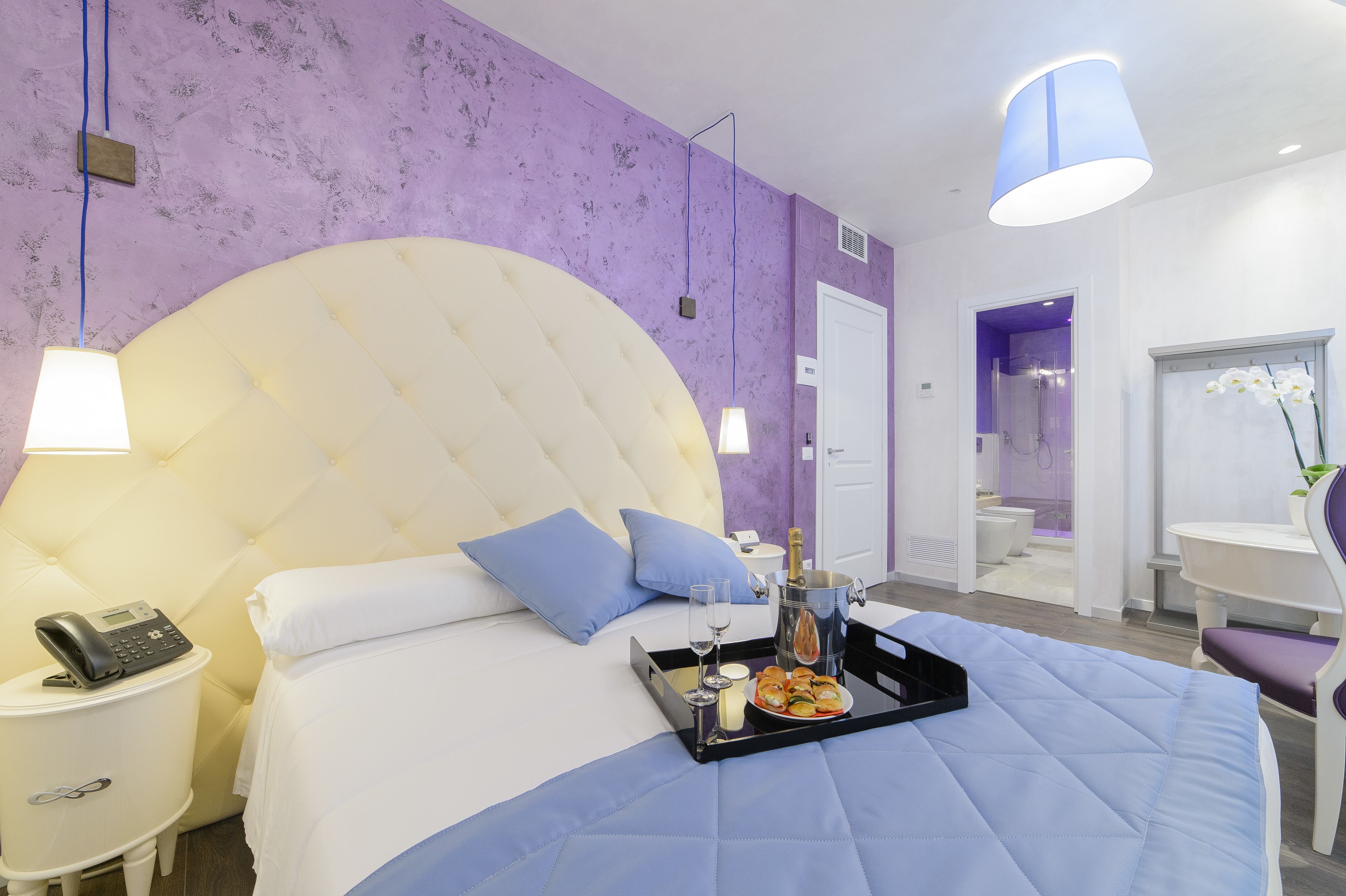 Une chambre d’hôtel moderne avec un grand lit, une table de chevet avec un téléphone, un plateau avec de la nourriture et des boissons, et une salle de bain visible à travers une porte ouverte.