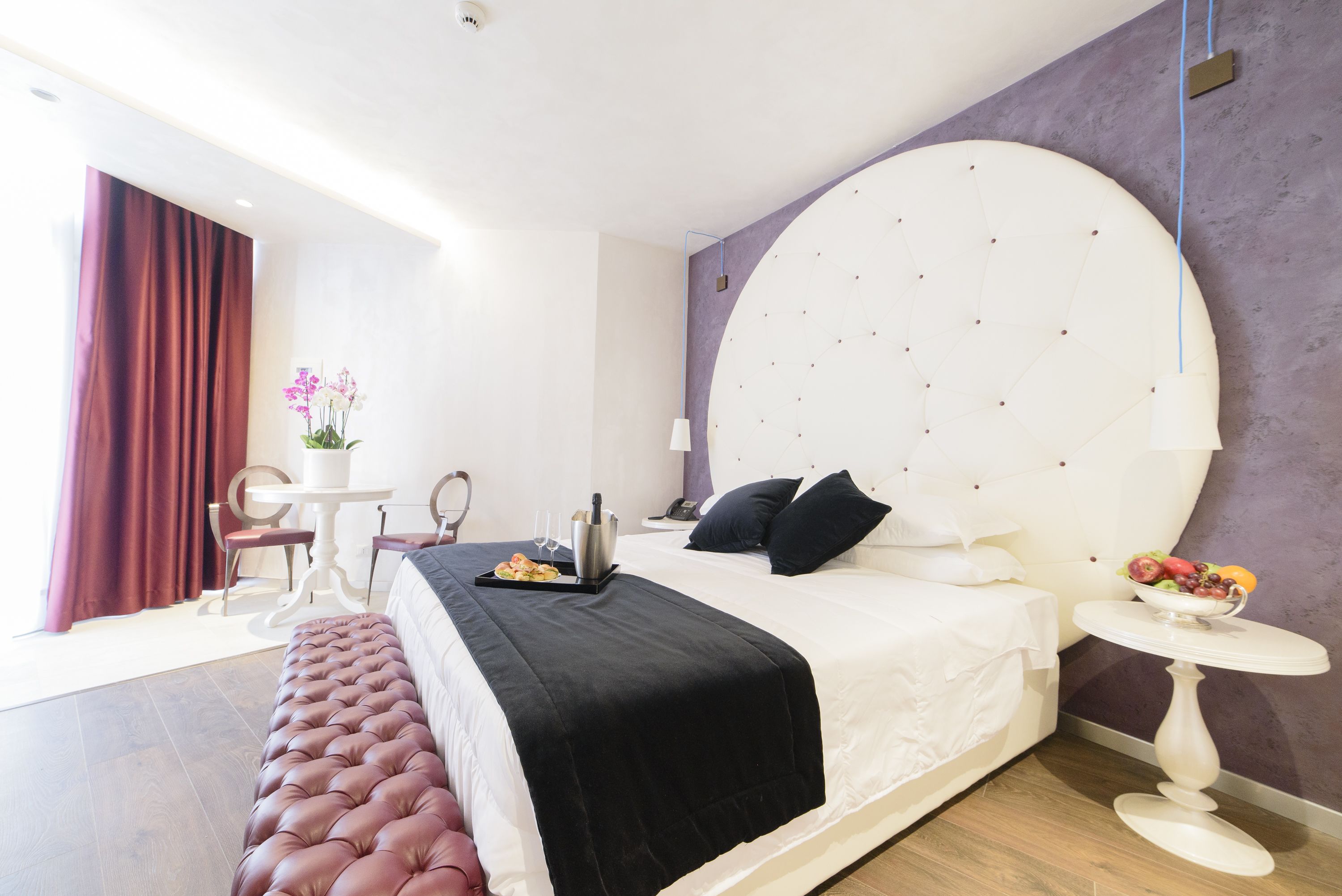 Una habitación de hotel moderna con una cama grande, un cabecero redondo, una pequeña mesa redonda con un cuenco de fruta, un banco y una mesa de comedor con sillas.