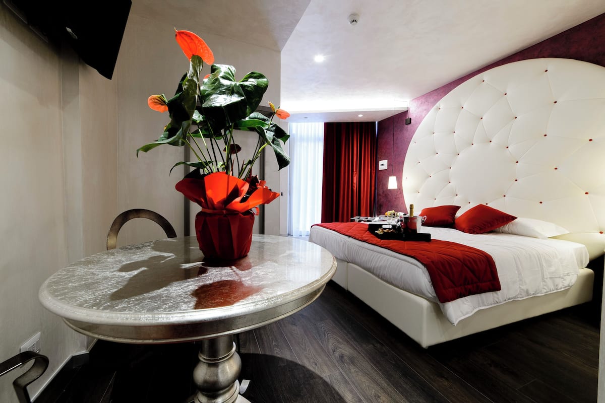 A modern hotel room with a round dining table, a large bed with a tufted headboard, and a red flower arrangement.