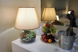 A table with a lamp, a fruit platter, a bottle of champagne, and two glasses.