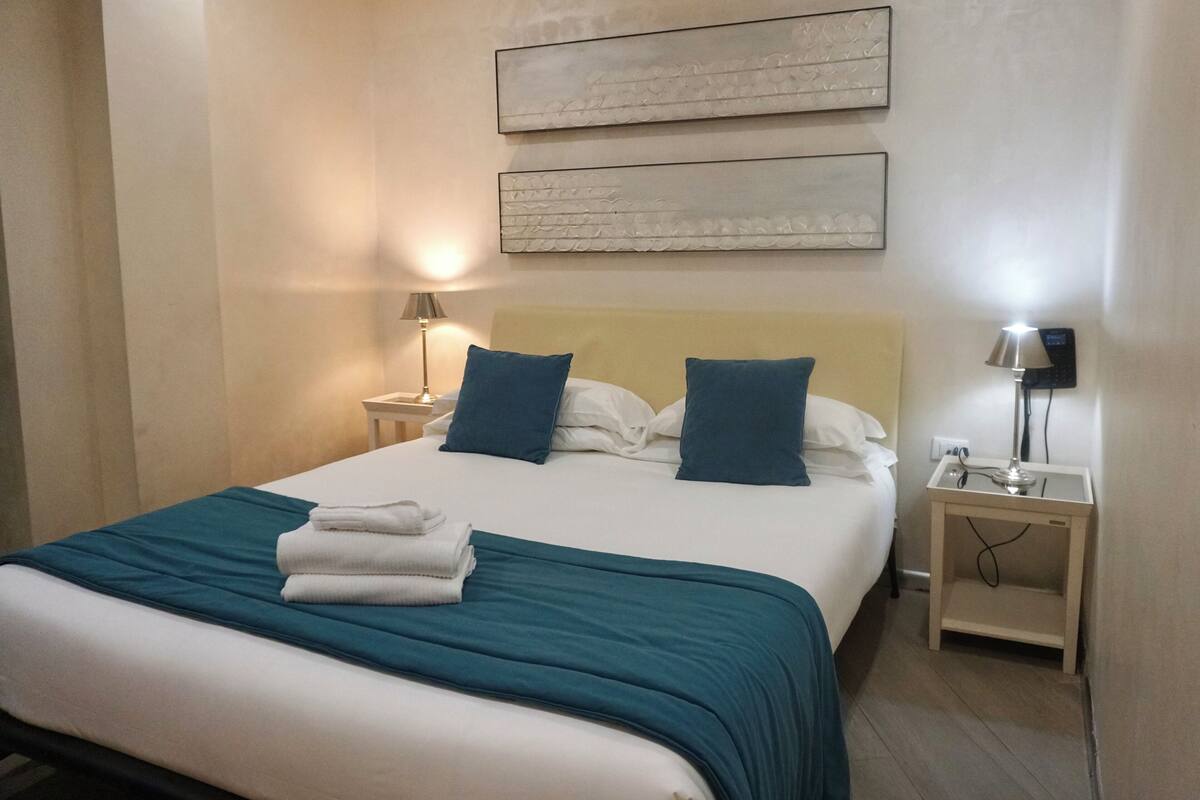 A neatly made bed with a teal bedspread and two pillows, a bedside table with a lamp, and a wall-mounted artwork above the bed.