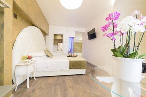 A modern bedroom with a large bed, a bedside table with fruit, a glass table with a vase of flowers, and a TV on the wall.
