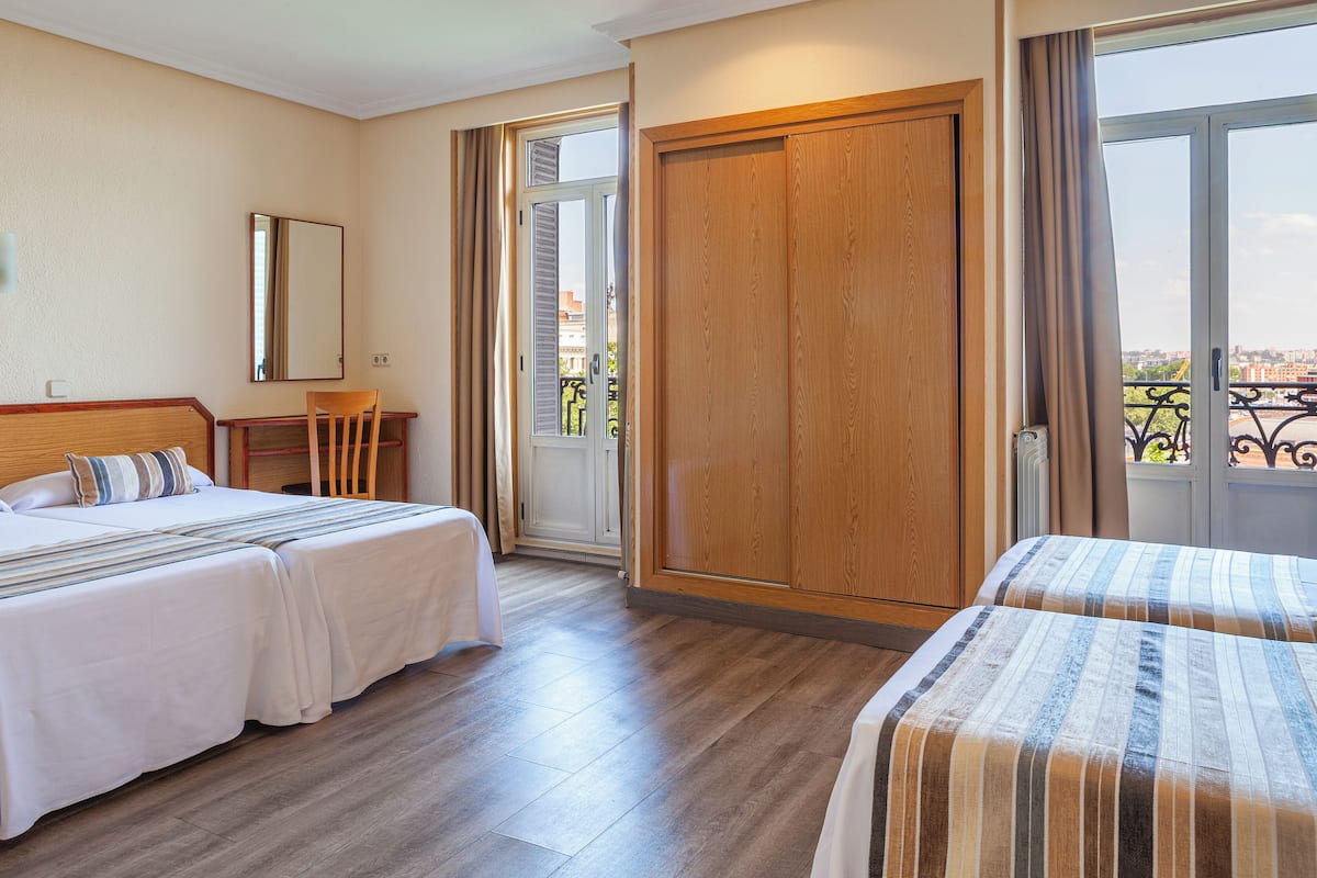 A hotel room with two beds, a wooden wardrobe, a small desk with a chair, a window with a view of buildings, and a balcony with a railing.