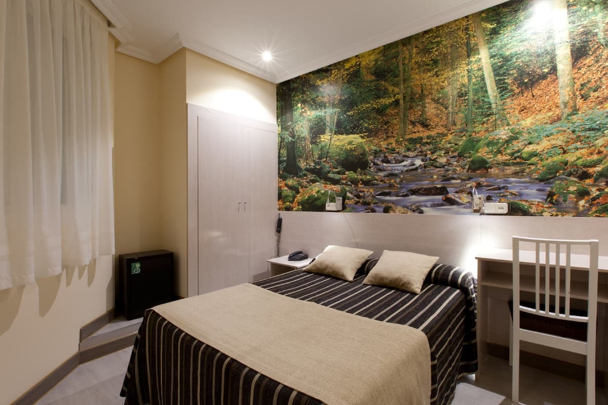 A bedroom with a bed, a desk, a chair, a wardrobe, and a wall mural of a forest scene.
