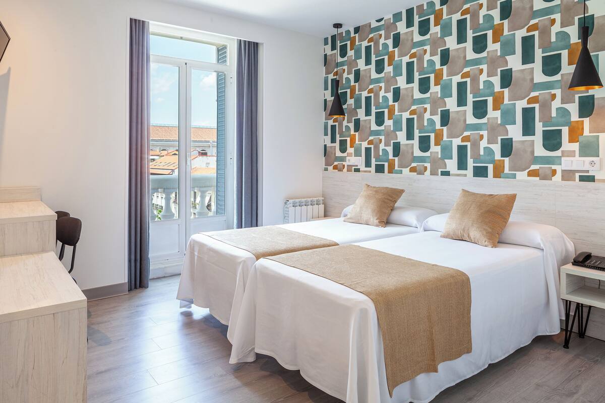A hotel room with two beds, a view of the city, a patterned wall, and a desk with a phone.