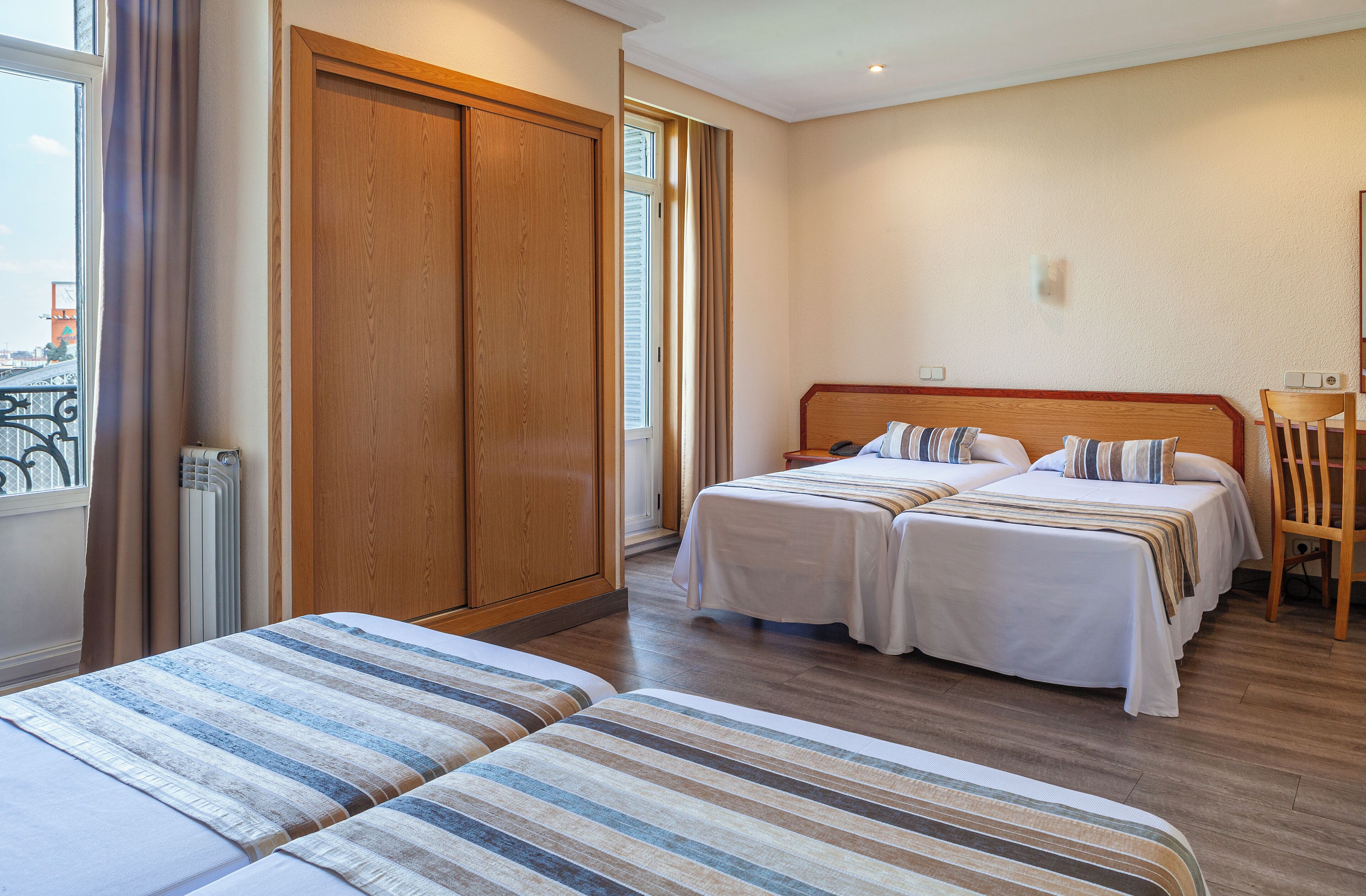 A hotel room with two beds, a wooden wardrobe, a radiator, and a window with curtains.