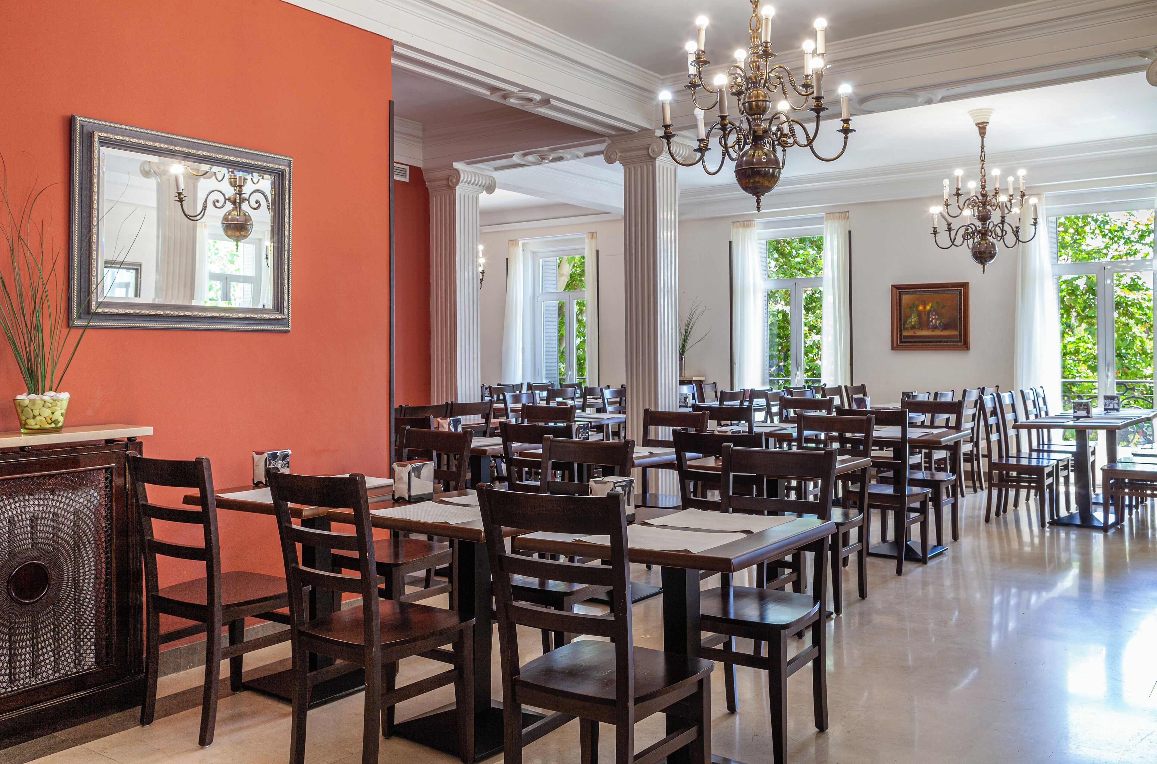 Un vaste espace dédié à la salle à manger, avec de nombreuses tables et chaises, de grandes fenêtres laissant entrer la lumière naturelle et un lustre.