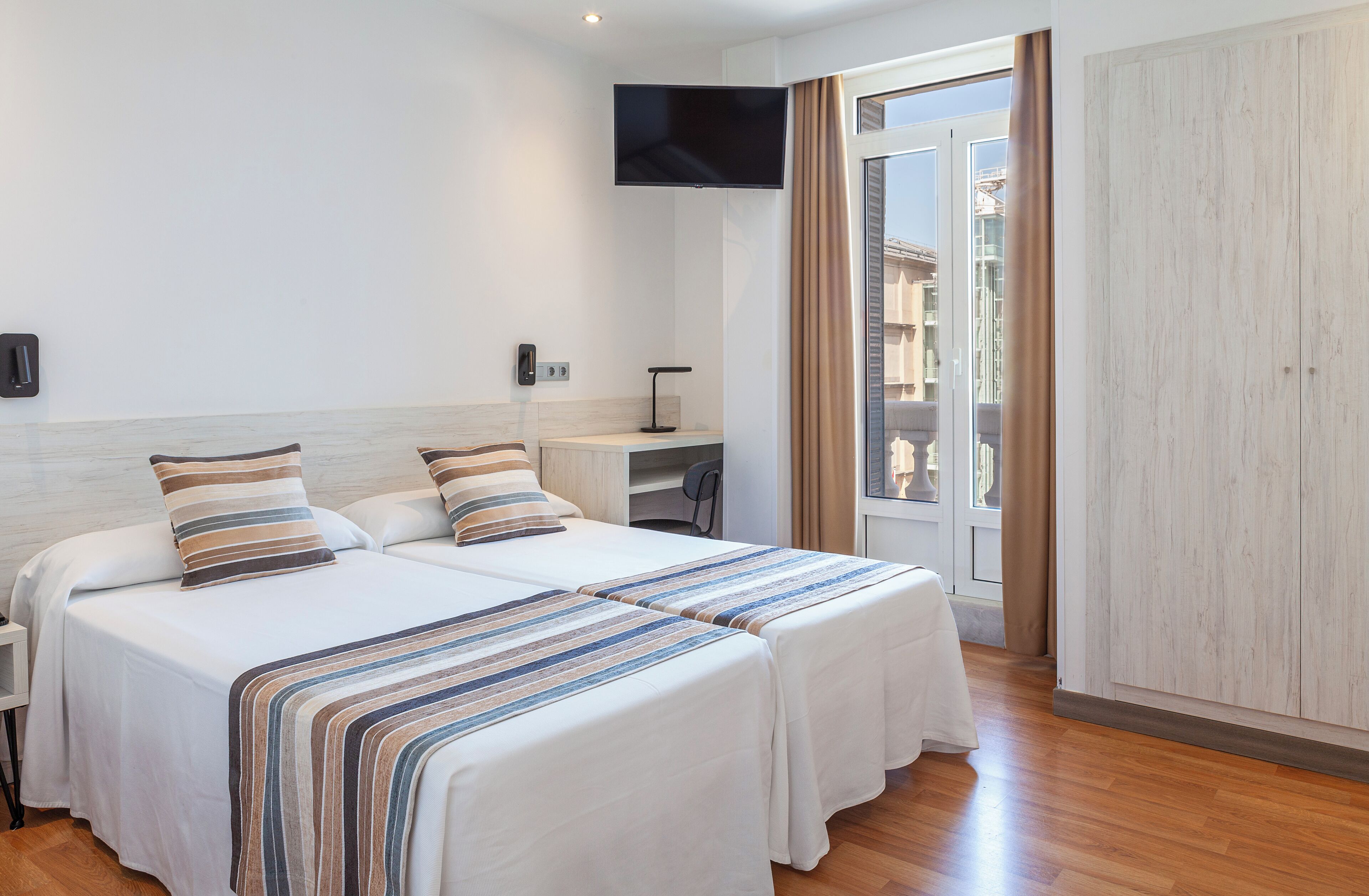 A hotel room with two beds, a TV, a desk, and a view of the city.