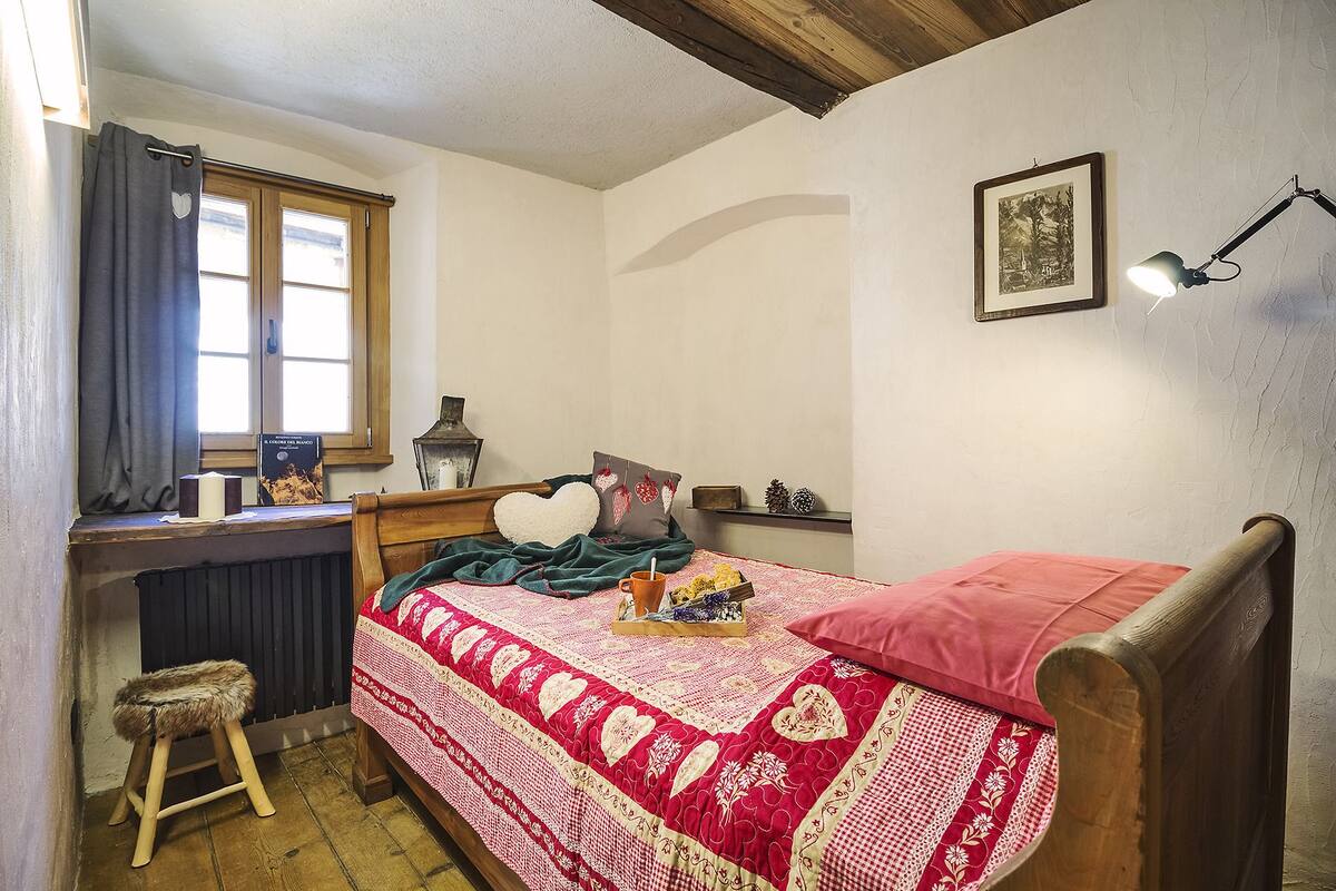 A cozy bedroom with a wooden bed, a small stool, a window with curtains, a bedside table with a lamp, and a framed picture on the wall.