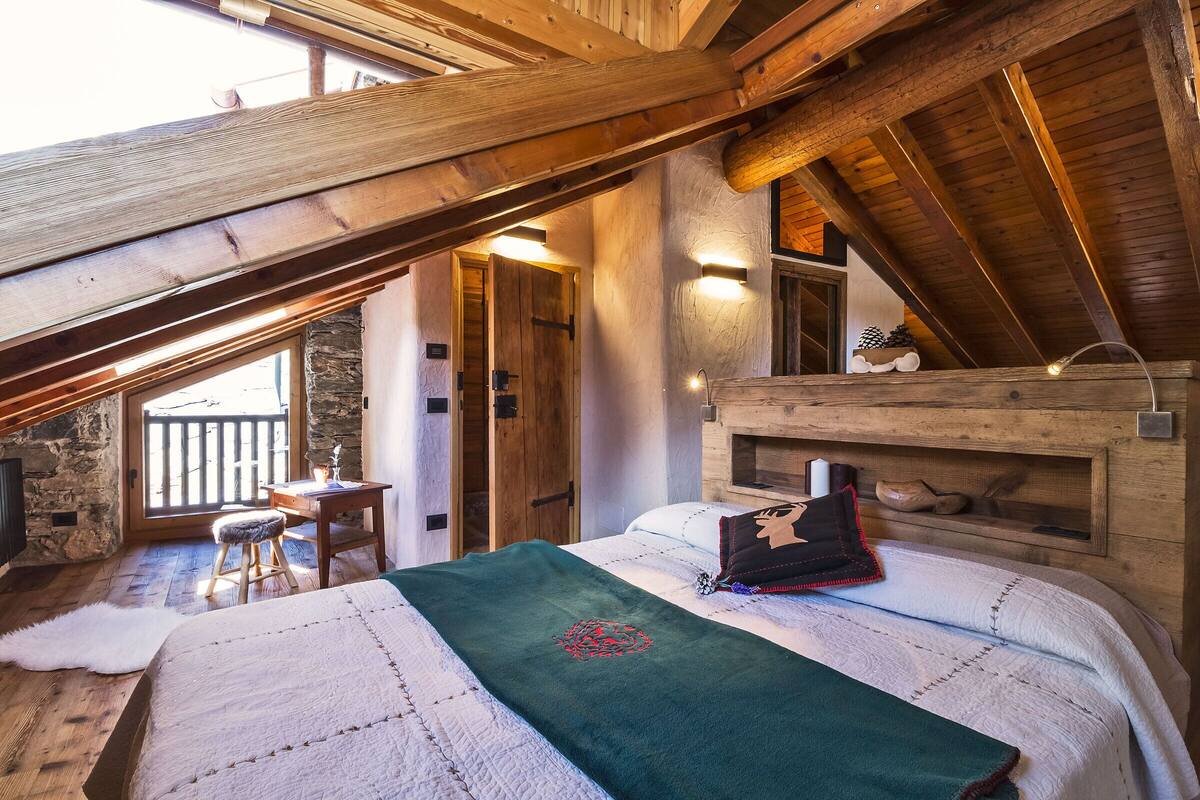 A cozy attic bedroom with a wooden ceiling, a bed with a green throw, and a stone wall.