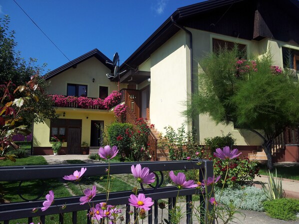 Una casa de dos pisos con un balcón decorado con flores rosas, un jardín con flores moradas y una valla de madera.