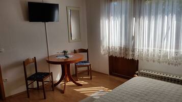 A room with a bed, a table with chairs, a TV mounted on the wall, and a window with sheer curtains.