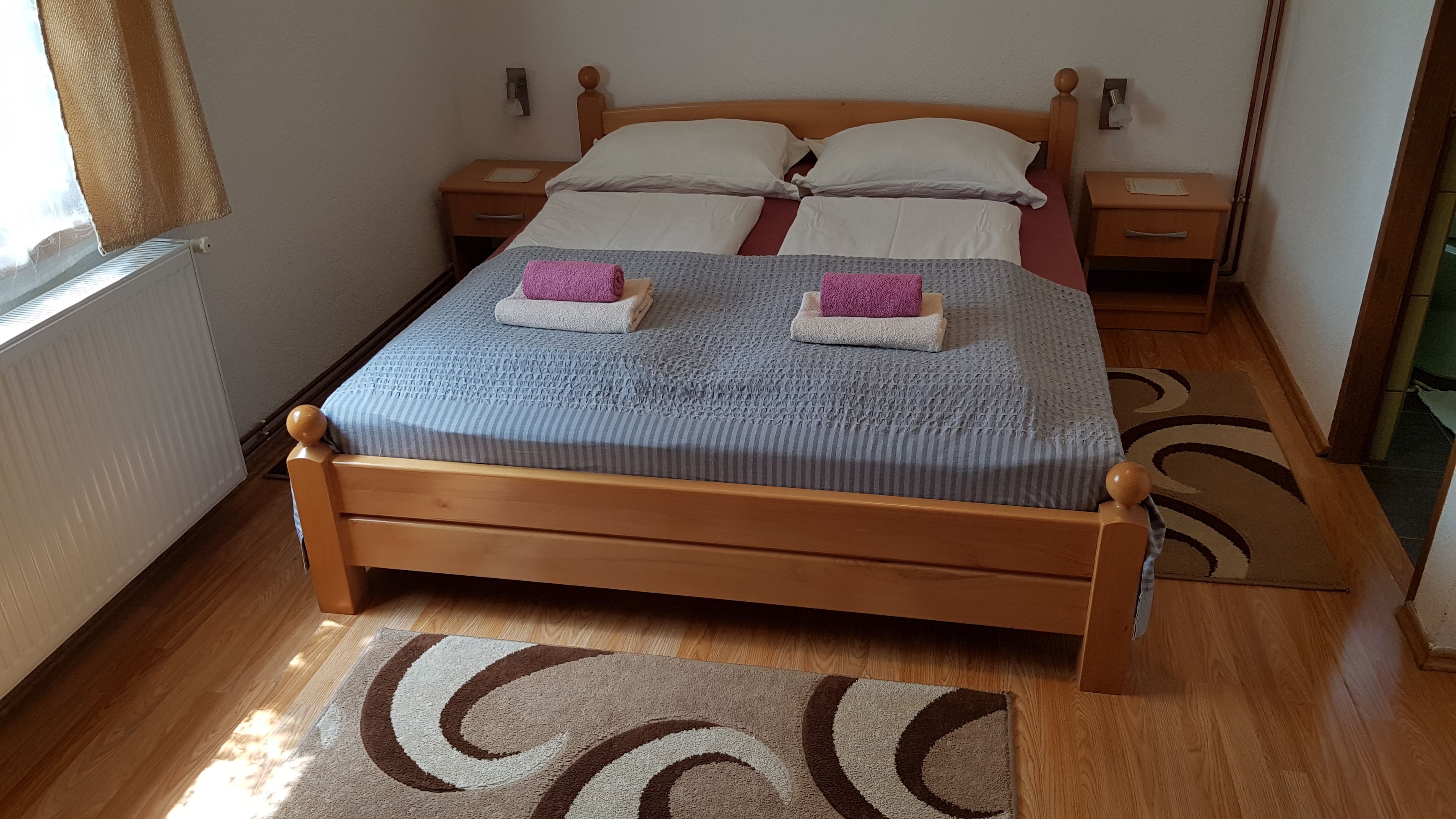 A neatly made bed with a striped blanket and two pillows, a wooden nightstand with a lamp, and a brown and white patterned rug on a wooden floor.