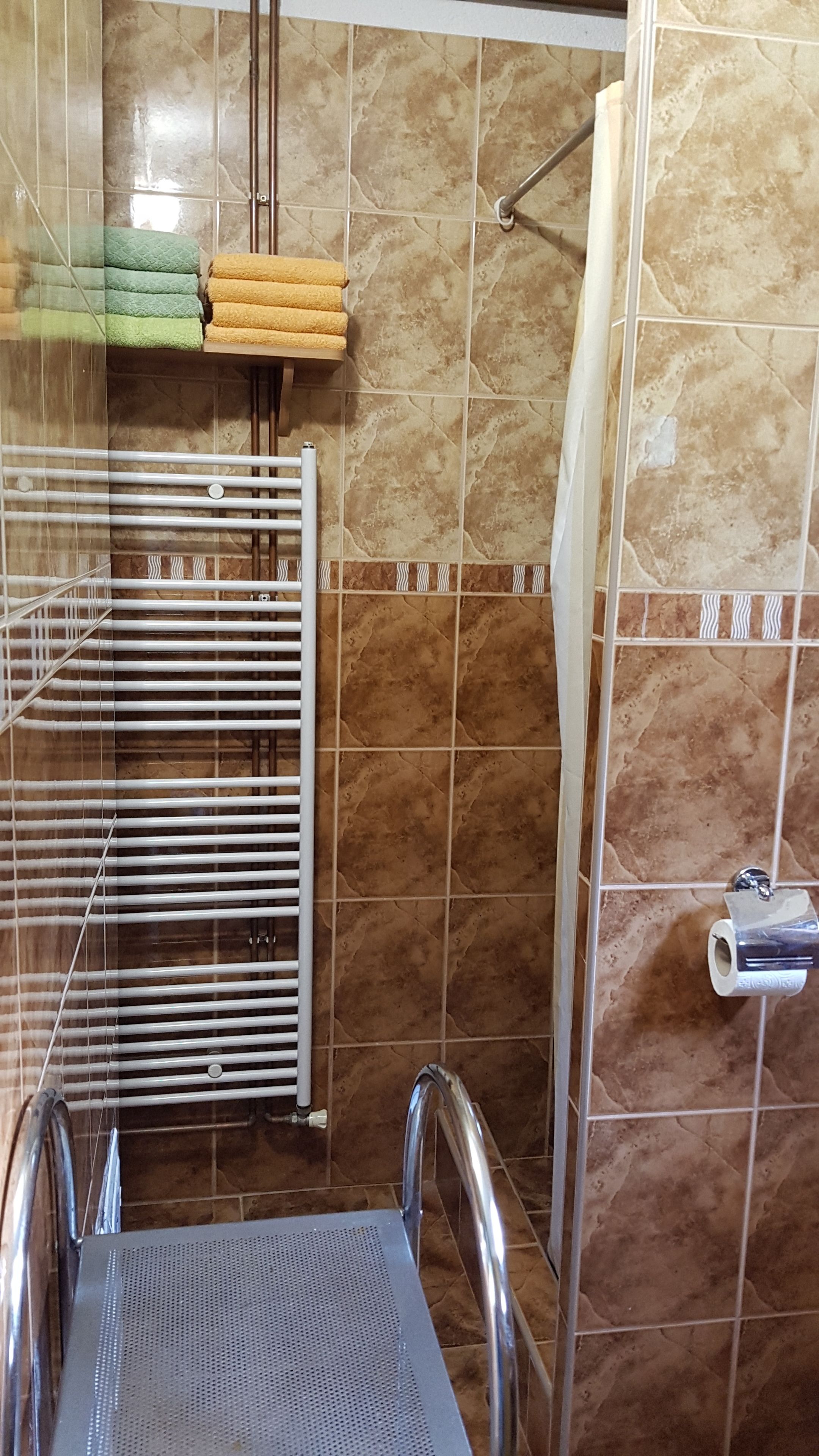 A bathroom with a shower area, a towel rack with folded towels, a radiator, and a toilet paper holder.