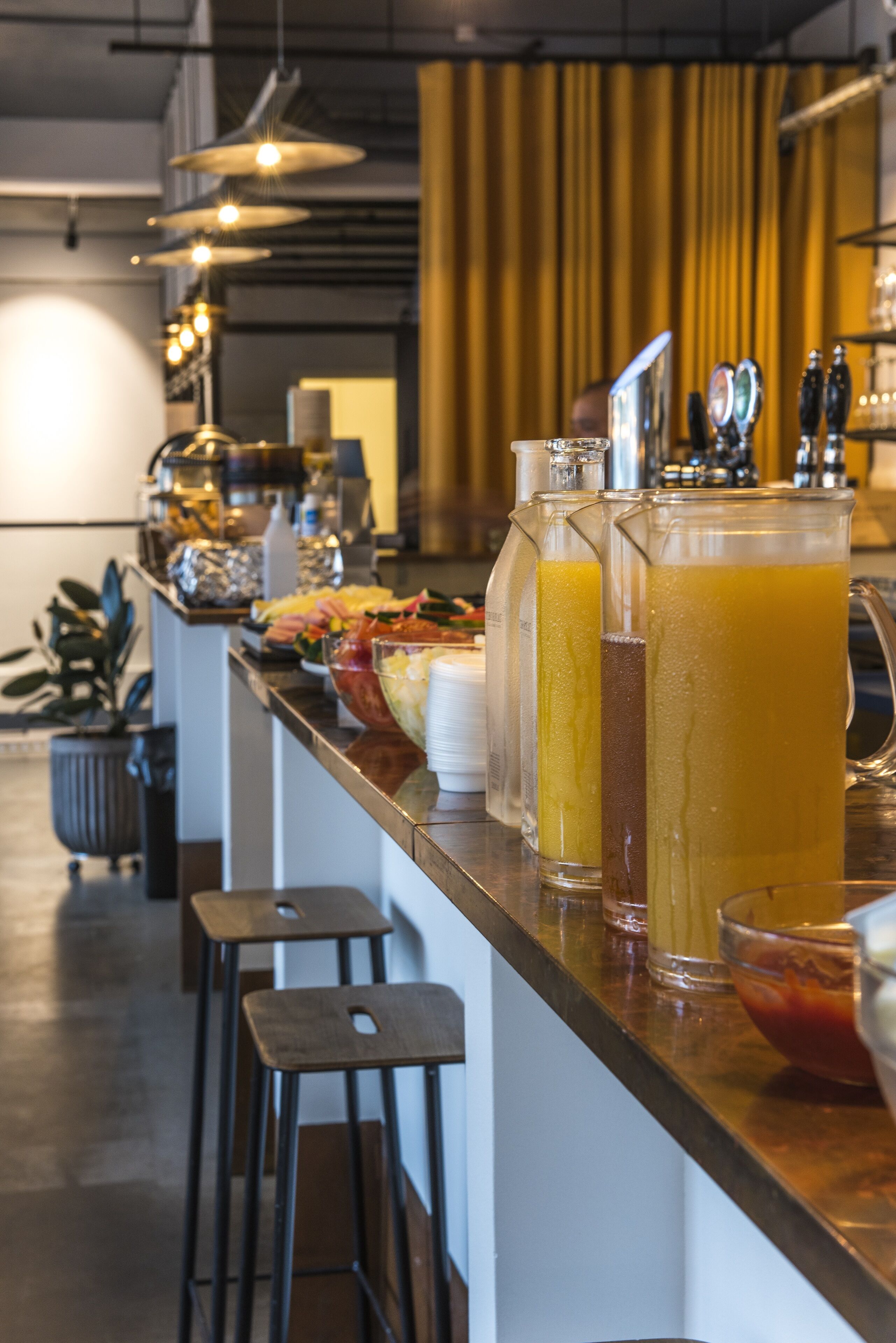 Un espace bar propose une variété de boissons, notamment une grande carafe de jus d’orange, un grand verre de thé glacé et un verre d’eau. On y trouve des tabourets et un comptoir derrière lequel se tient un barista.