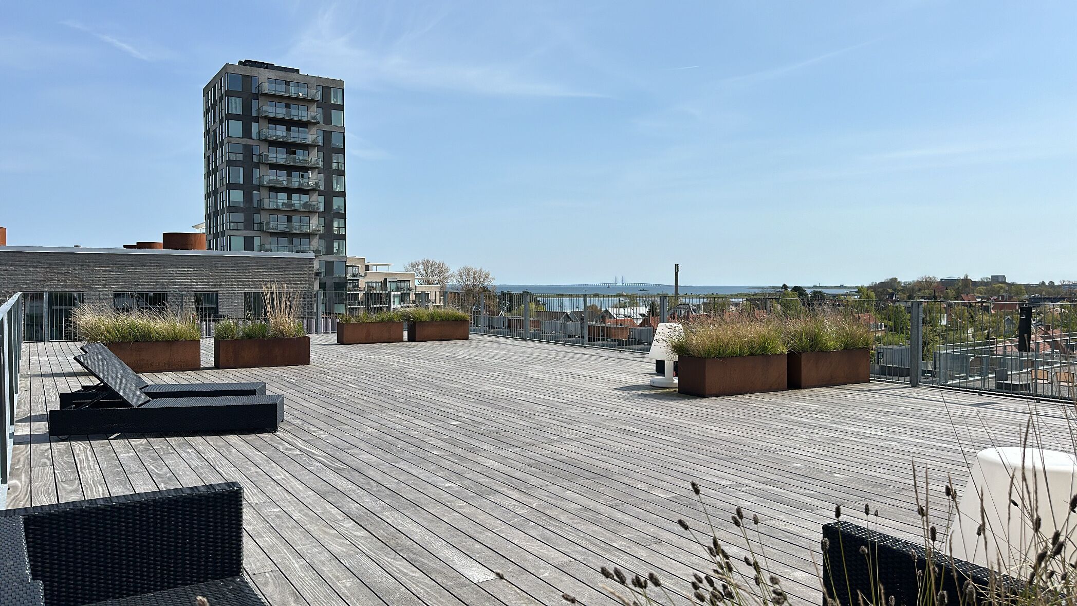 Een dakterras met houten terrassen, plantenbakken met groen en uitzicht op zee en gebouwen.