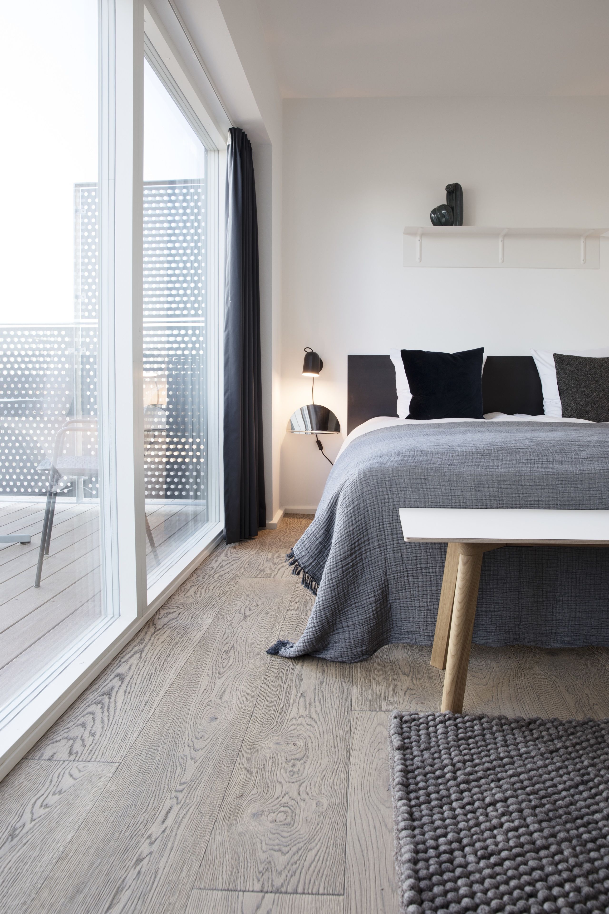 A modern bedroom with a large window, a bed with a grey blanket, a bedside table, and a wooden floor.