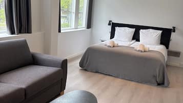 Una habitación de hotel moderna con un sofá, una cama con una manta gris, una mesita redonda con un control remoto y una ventana con cortinas.