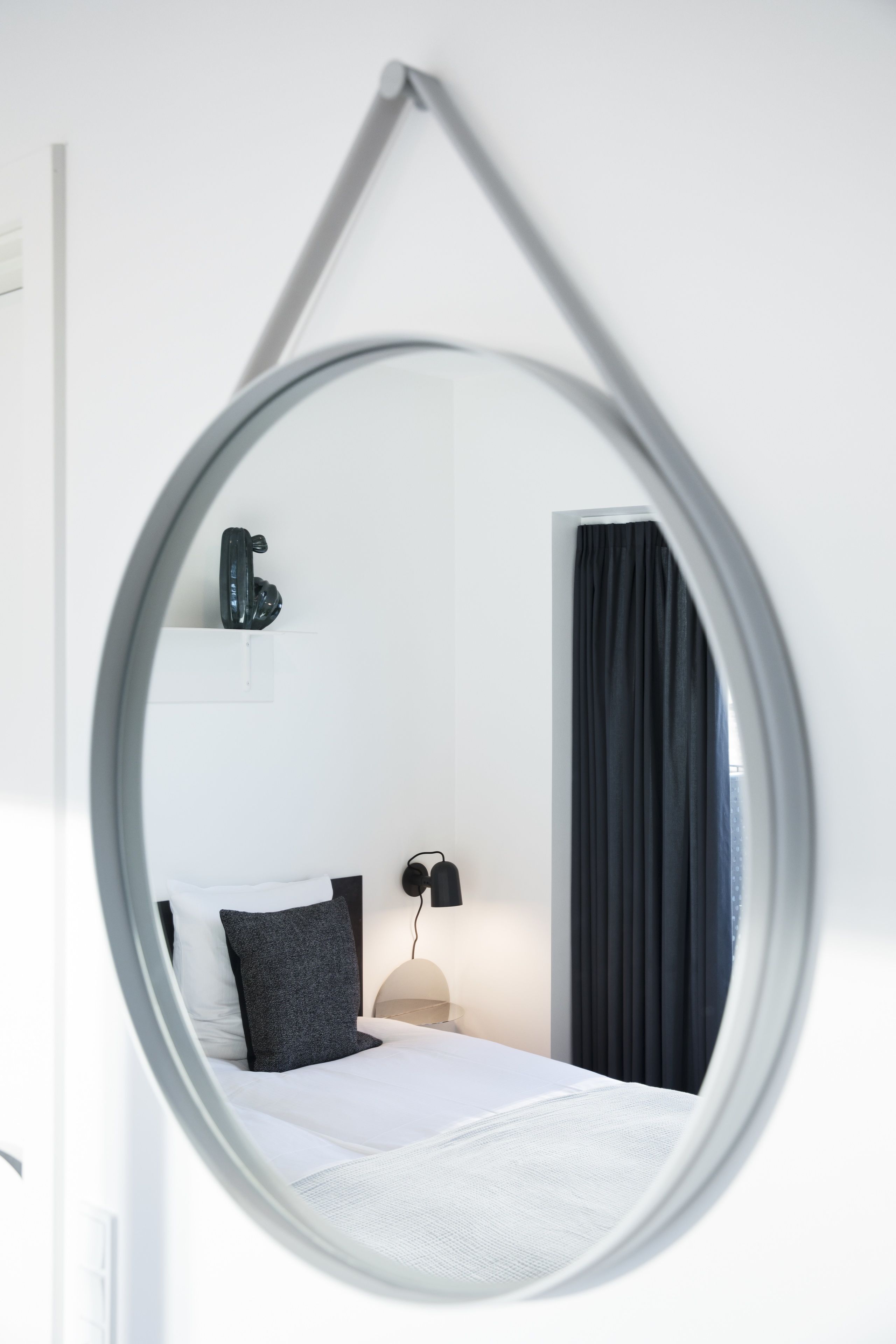 Una habitación de hotel moderna con una cama, un espejo redondo, una mesita de noche, una lámpara y una ventana con cortinas.