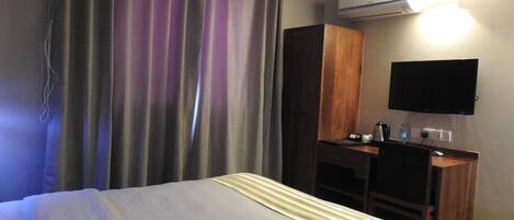 A hotel room with a bed, a wardrobe, a desk with a chair, a flat-screen TV, and an air conditioning unit.