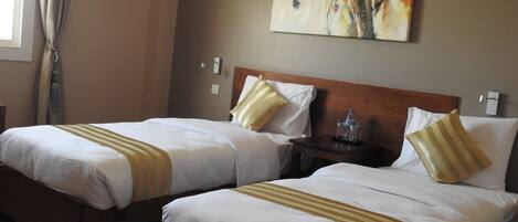 Habitación de hotel con dos camas, cada una con ropa blanca y almohadas a rayas, un cabecero de madera, una mesita de noche con una botella de agua y un cuadro en la pared.