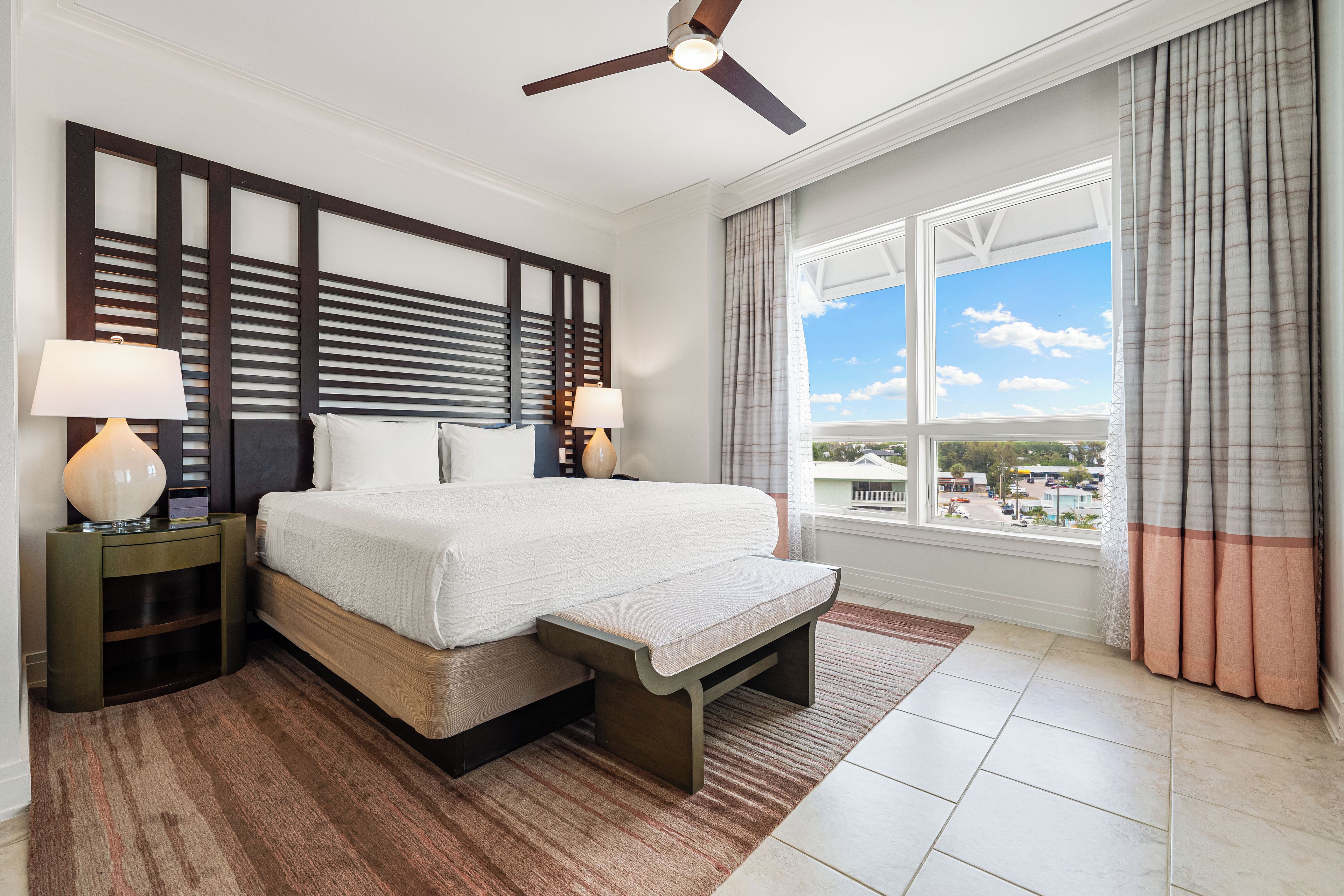 A modern hotel room with a large bed, two bedside lamps, a ceiling fan, and a view of the city through large windows.